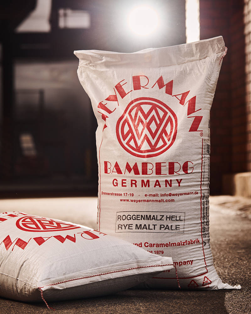 Two sacks of rye malt are next to each other, one lying on the floor and the other propped up. Dramatic spotlights shine into the camera and onto the malt bags. 